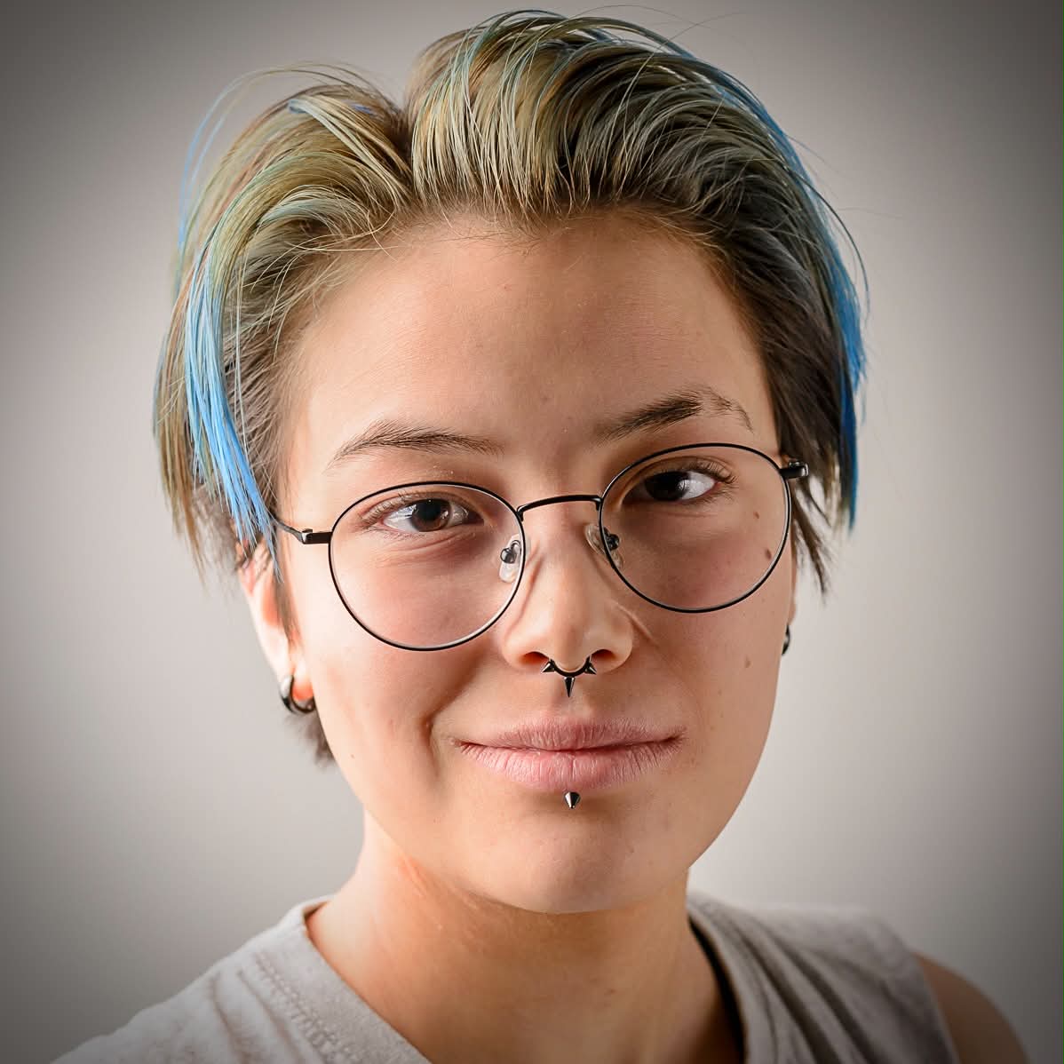 close up picture taken from the front.
                        The person on the camera is wearing glasses and have a few piercings as well as dyed blue hair.
                        their eyes are brown and they are wearing a grey shirt